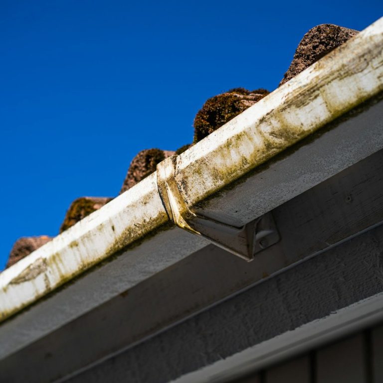 Nahaufnahme einer Dachrinne mit Verfärbungen und blauem Himmel im Hintergrund.