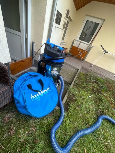 Blauer Staubsauger mit Schlauch und Transporttasche vor einem Haus.