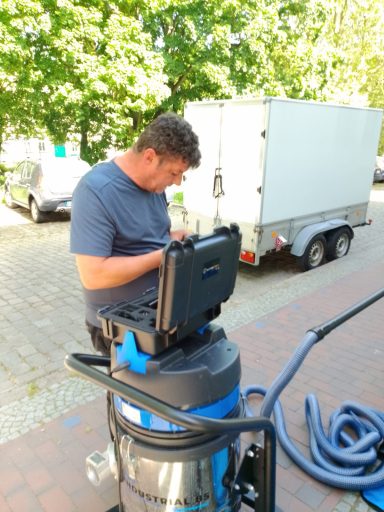 Mann sitzt auf einem Staubsauger und nutzt einen Laptop in einer Außenumgebung.